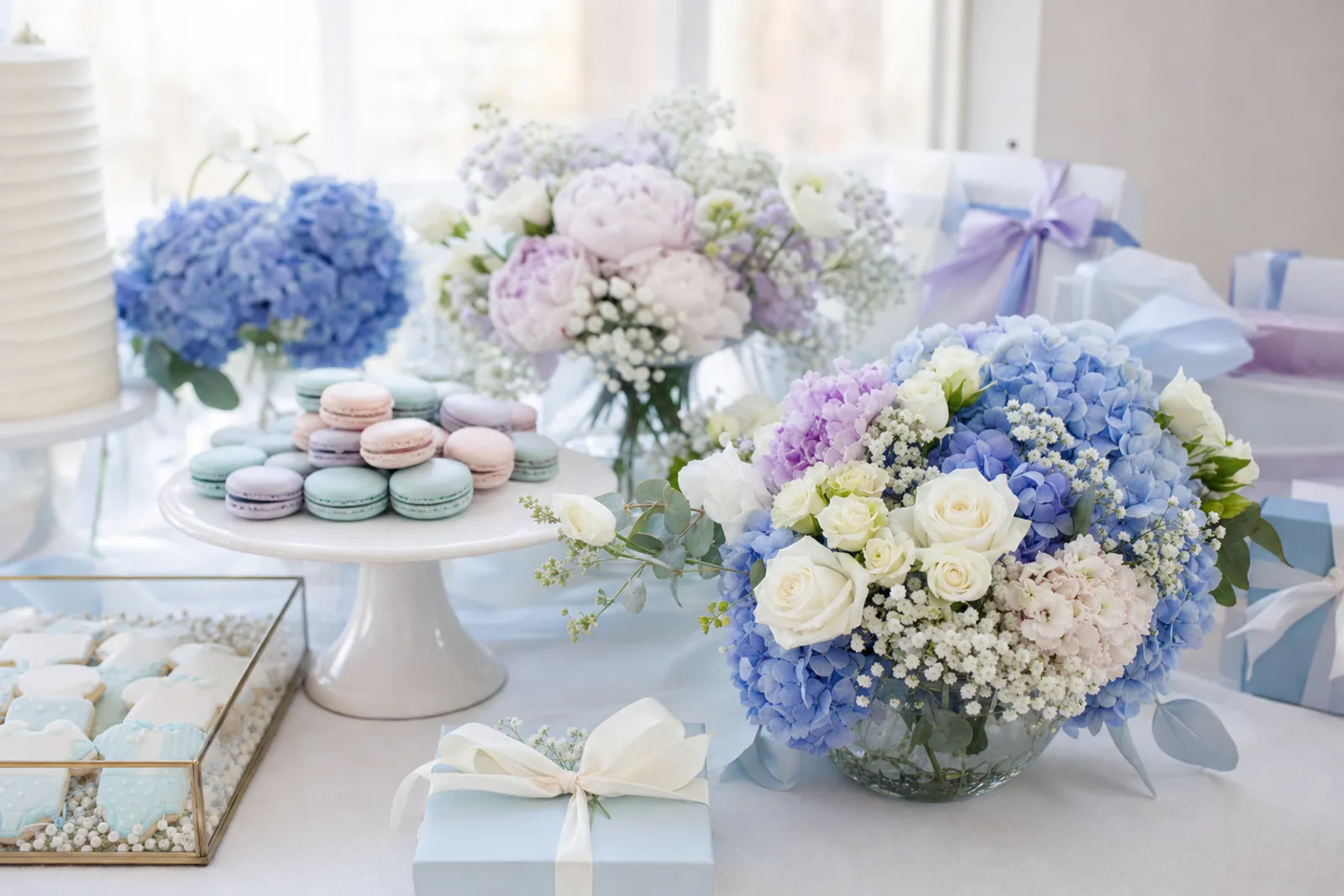 Pastel baby shower table with a low bowl of blue hydrangea and white roses, a vase of pale pink and lilac peonies with baby's breath, macarons and gifts tied with ribbon