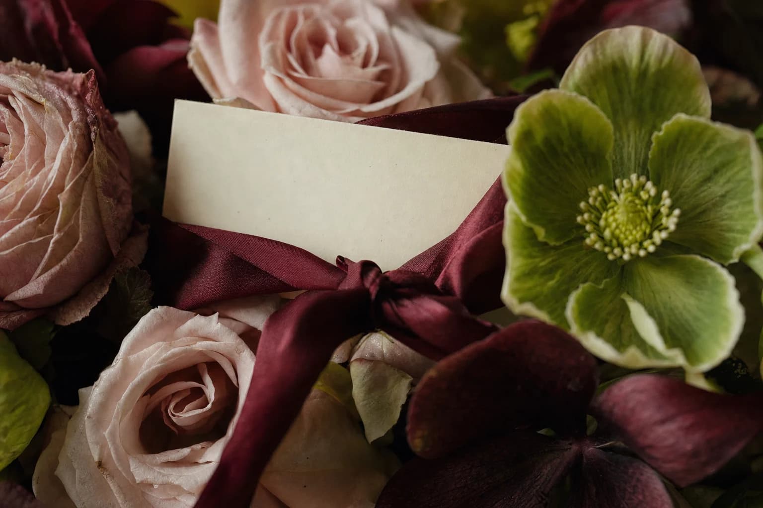 A blank gift card tied into a bouquet of blush roses and green hellebores with garnet ribbon